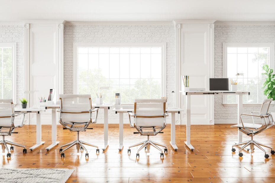 Un espace de bureau lumineux et moderne avec des bureaux blancs, des chaises ergonomiques et de grandes fenêtres laissant entrer la lumière naturelle. La pièce présente des murs de briques blanches, un sol en bois et un poste de travail debout.