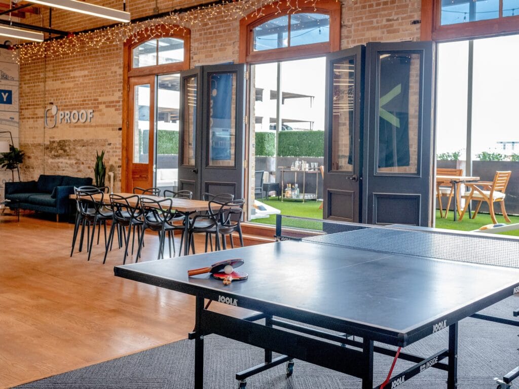 Espace de détente en bureau ouvert avec table de ping-pong, coin salon et terrasse extérieure avec mobilier de jardin.